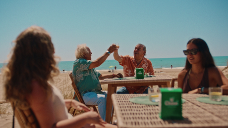 Dos hombres mayores brindando en una mesa de terraza en la playa, con servilleteros verdes y otras personas alrededor.