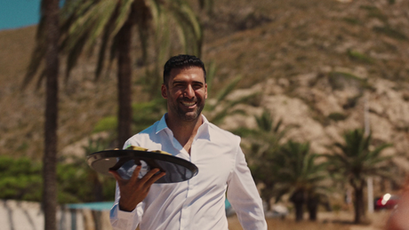 Camarero sonriente con camisa blanca sujetando una bandeja con platos al aire libre junto a palmeras y montañas.