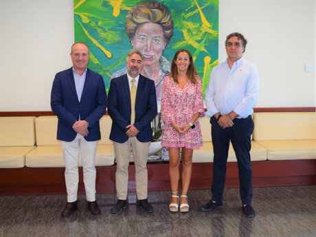 Cuatro personas vestidas de forma formal posan de pie frente a un cuadro colorido en un vestíbulo con bancos.