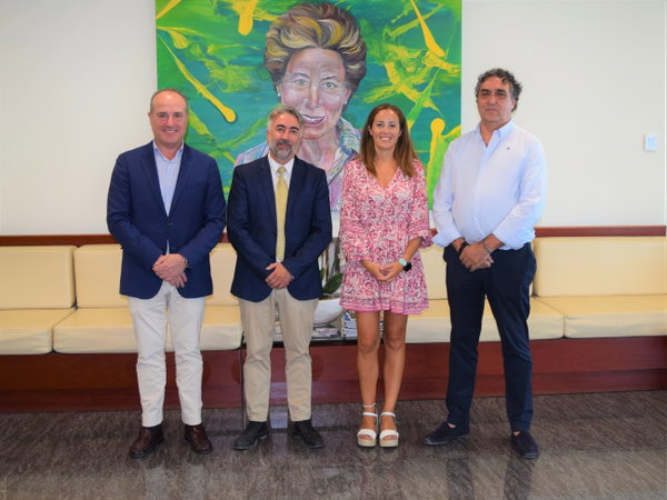 Cuatro personas vestidas de forma formal posan de pie frente a un cuadro colorido en un vestíbulo con bancos.