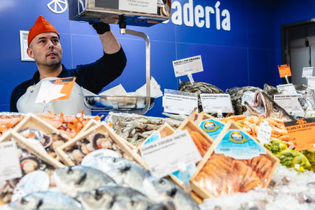 Mostrador de pescadería con sardinas y mariscos sobre hielo y un pescadero con gorro naranja pesando producto en una báscula.