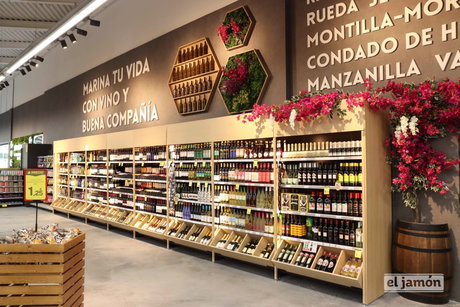 Lineal de vinos en supermercado con botellas, estanterías de madera y decoración floral; logo el jamón visible en esquina.