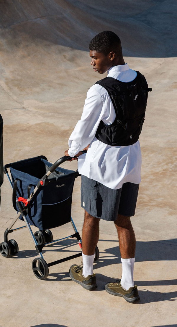 Joven empuja un cochecito de paseo azul, viste camisa blanca, chaleco negro, bermudas oscuras y zapatillas verdes.