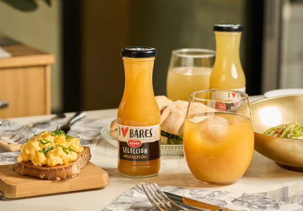 Botella de zumo Juver con etiqueta I Love Bares selección melocotón, vaso con hielo, tostada y platos de desayuno.