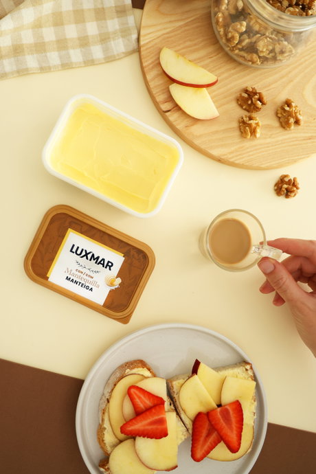 Tarrina de mantequilla Luxmar abierta junto a tostadas con rodajas de manzana y fresas, nueces y mano sujetando una taza de c