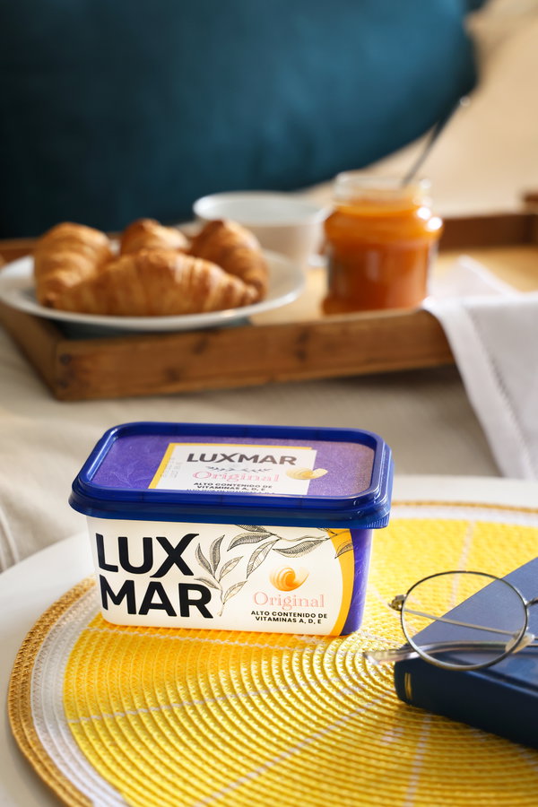 Tarrina de margarina LUXMAR Original sobre un mantel amarillo, con croissants y un tarro de mermelada al fondo.
