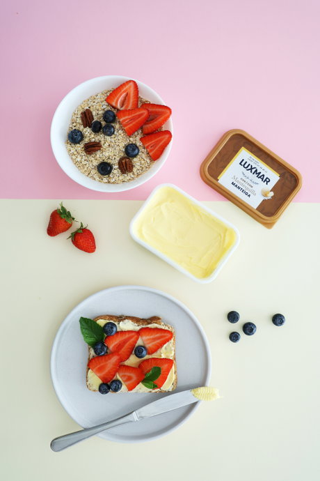Tostada con mantequilla, fresas y arándanos sobre un plato; al lado bol de avena y tarrina Luxmar de mantequilla.
