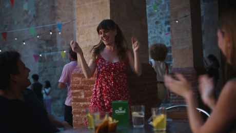 Mujer joven bailando con vestido rojo de flores junto a mesa con vasos y un envase verde Chovi en una fiesta al aire libre.