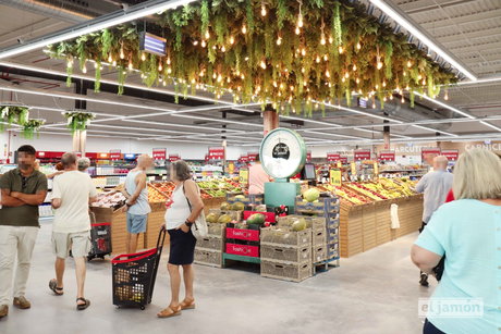 Sección de frutas y verduras en supermercado El Jamón con báscula antigua, cajas de melones y clientes comprando.
