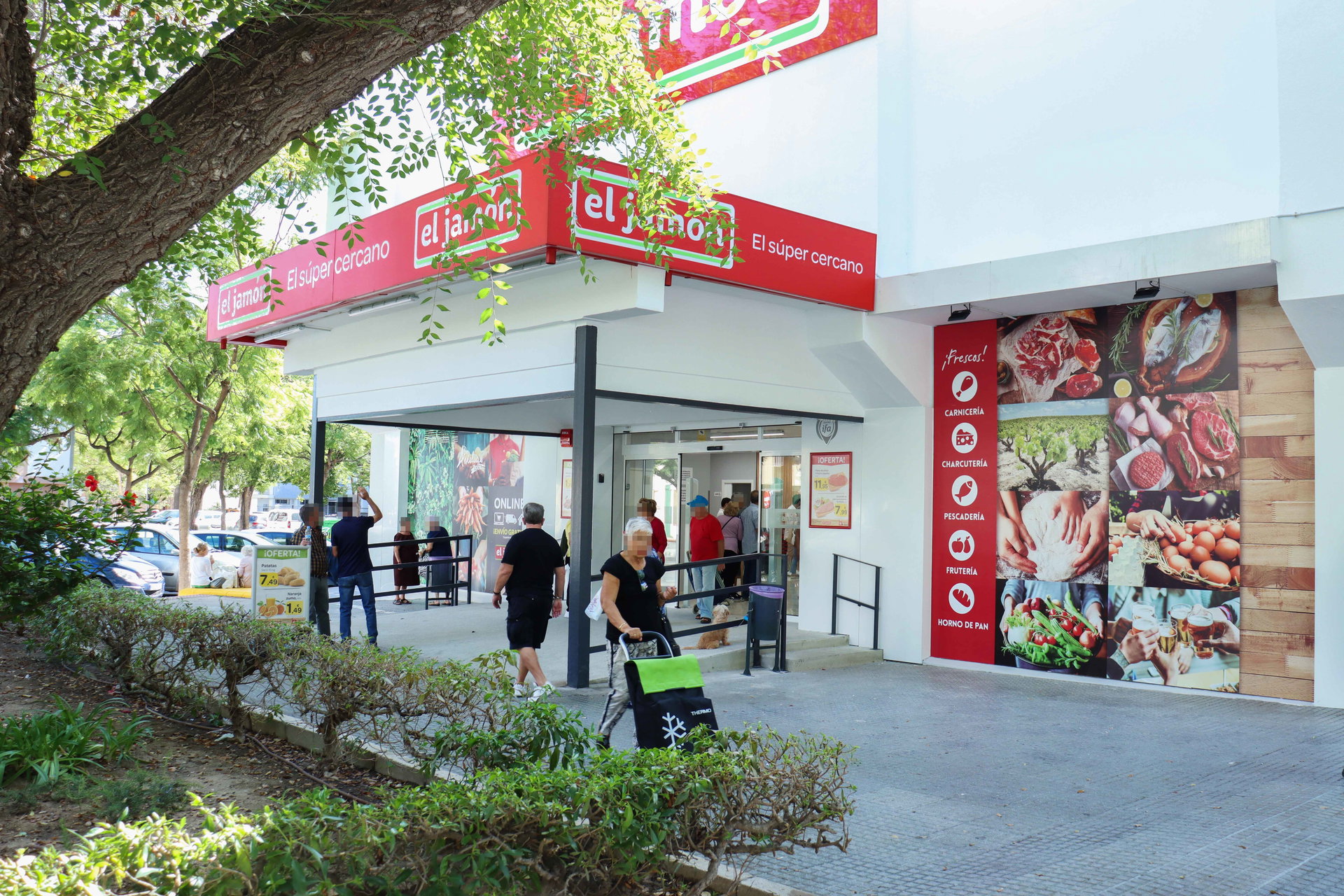 Fachada y entrada del supermercado Supermercados El Jamón con clientes entrando y carro de la compra.