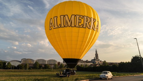 Globo aerostático amarillo con el logo ALIMERKA inflado en un campo, cesta en tierra y coche clásico aparcado.