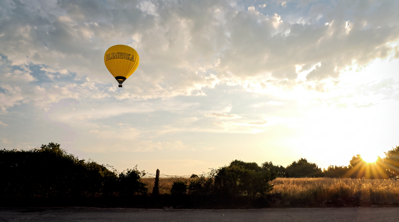 Globo aerostático amarillo con el logo ALIMERKA sobre un paisaje de campo al atardecer con el sol bajo.