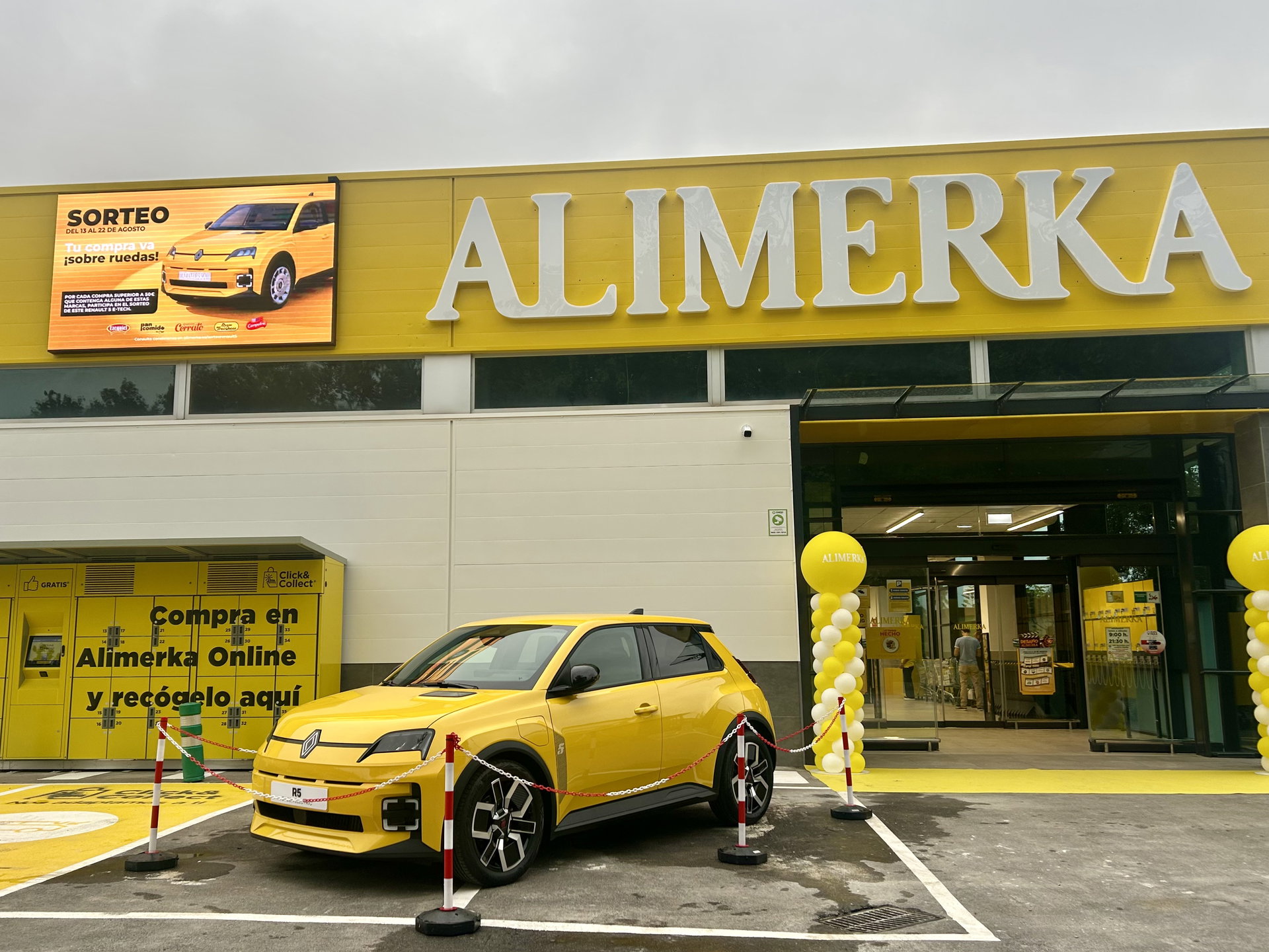 Coche promocional amarillo aparcado frente a la fachada de ALIMERKA con columnas de globos y señalética de recogida online.