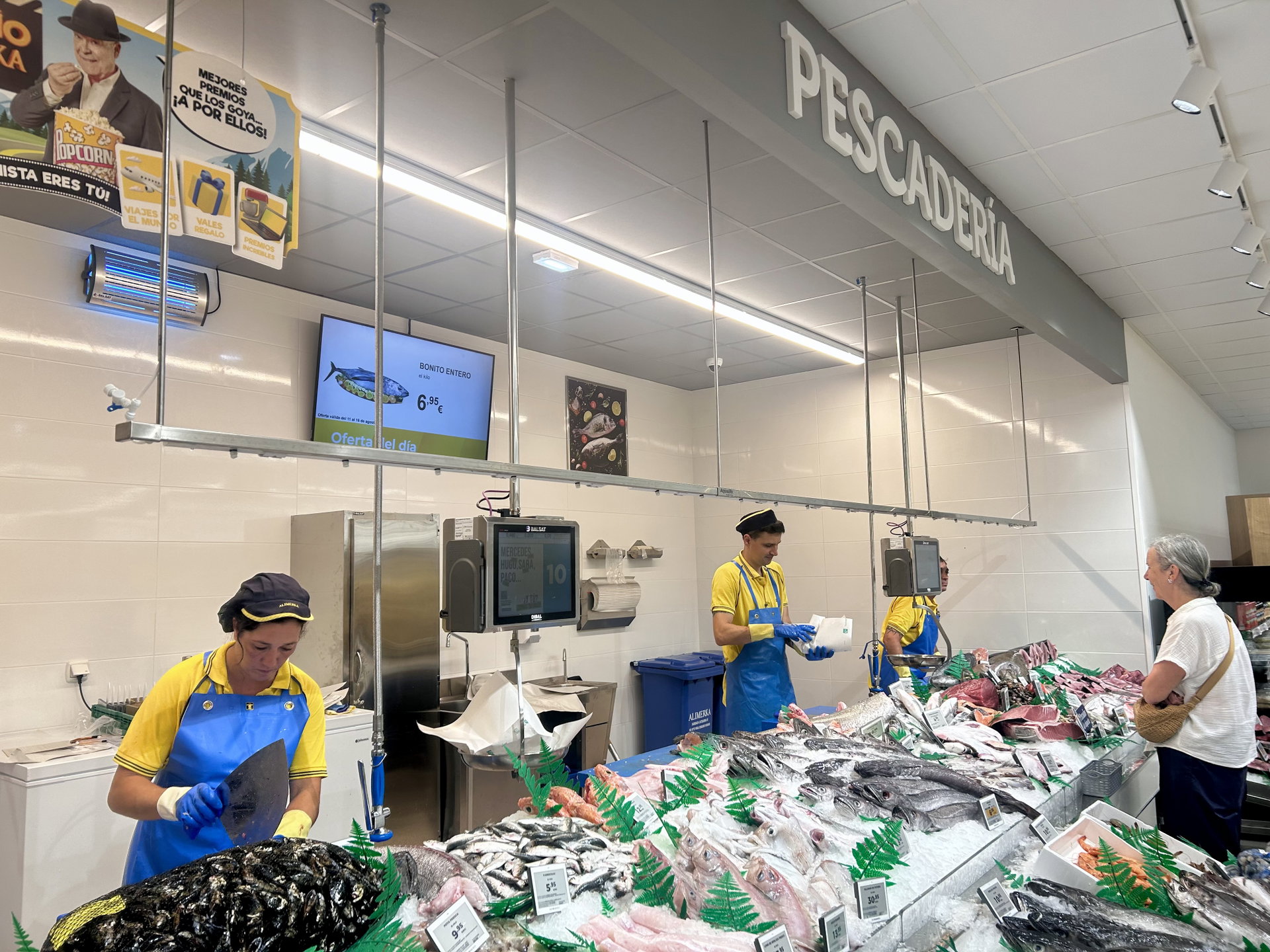 Mostrador de pescadería con variedad de pescados y mariscos sobre hielo y dos empleados con uniforme amarillo y azul atendien