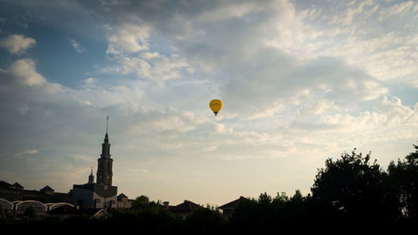 Globo aerostático amarillo sobre el skyline urbano al atardecer, con un campanario y árboles en primer plano.
