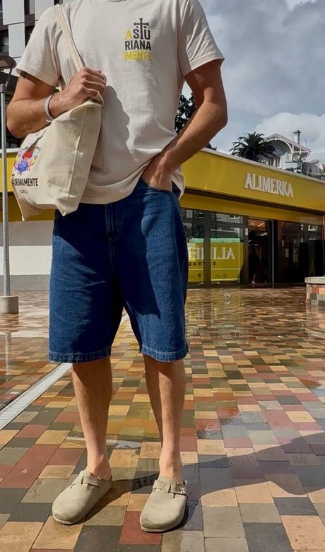 Hombre de pie con camiseta clara, bermudas vaqueras y zuecos beige, bolsa de tela al hombro y fachada de Alimerka al fondo.