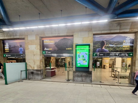 Vestíbulo de estación con cartelería de Alimerka, tres accesos y un panel digital publicitario verde.