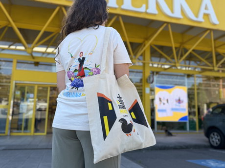 Mujer de espaldas con camiseta blanca ilustrada y bolso tote de tela con el texto ASTURIANA, frente a una fachada amarilla.