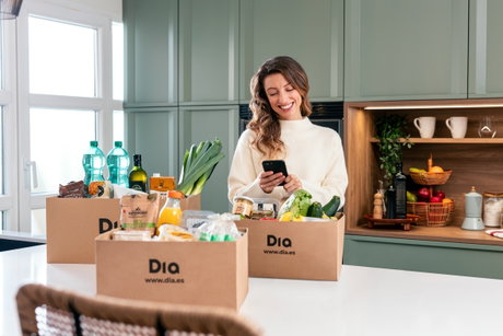 Cajas de supermercado Dia llenas de alimentos junto a una mujer sonriente que mira el móvil en la cocina.