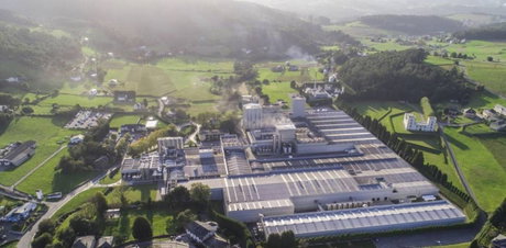 Planta industrial vista aérea con grandes naves y cubiertas con paneles solares, chimeneas y campos verdes alrededor.