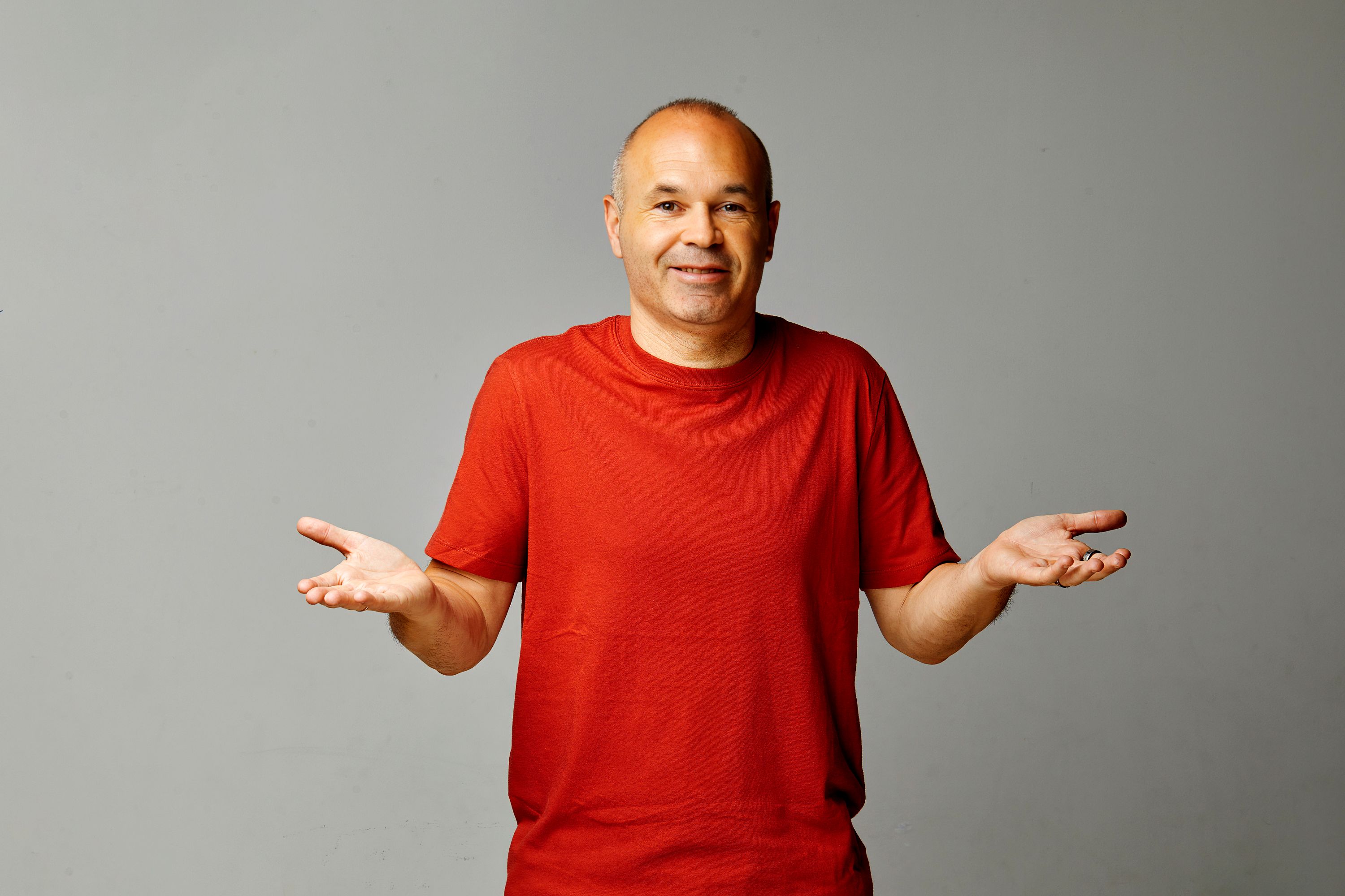 Hombre de mediana edad con camiseta roja, brazos abiertos y expresión de duda sobre fondo gris.