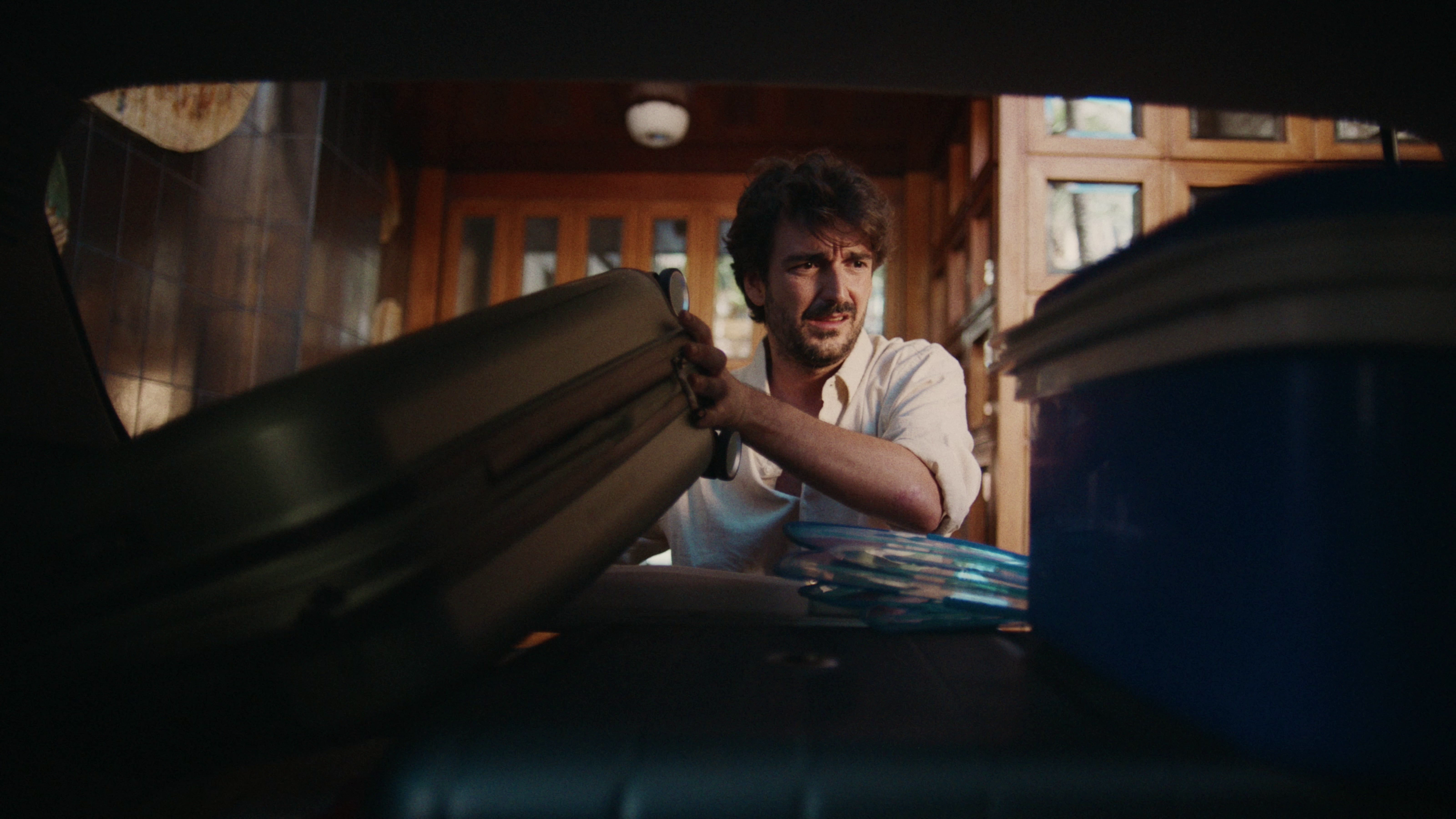 Hombre colocando una maleta grande en el maletero de un coche, junto a una nevera portátil azul y platos apilados.